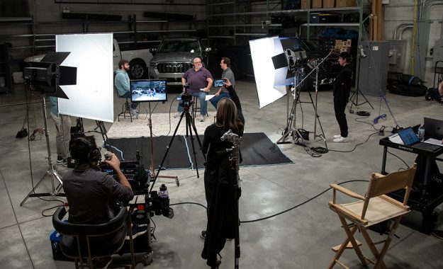 Production crew filming a video of three people talking in front of two trucks.